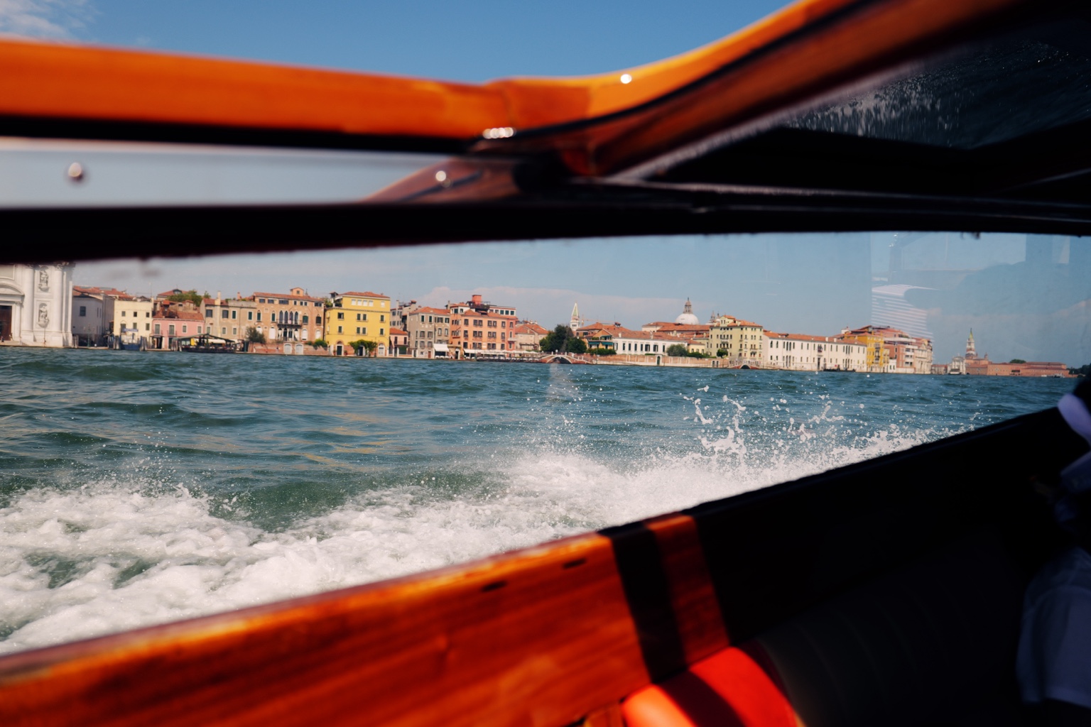 Blue water, breaking waves, and waterfront buildings seen through the orange wooden window frame of a boat.